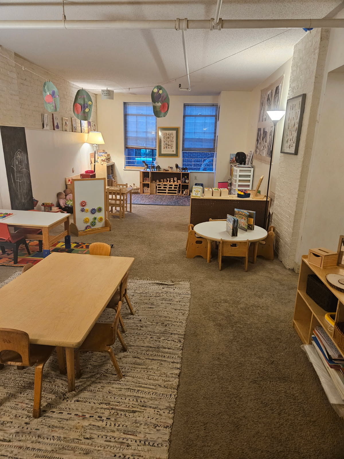 Wide view of the main room at Learning Sprouts: warm lamp light, children's artwork strung overhead, and wooden shelving and tables receding into depth.
