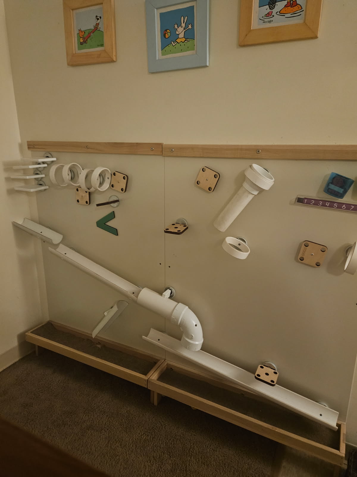 A wall-mounted ball run: wooden and white PVC ramp pieces, tubes and rings fixed to the wall above a wooden catch track.