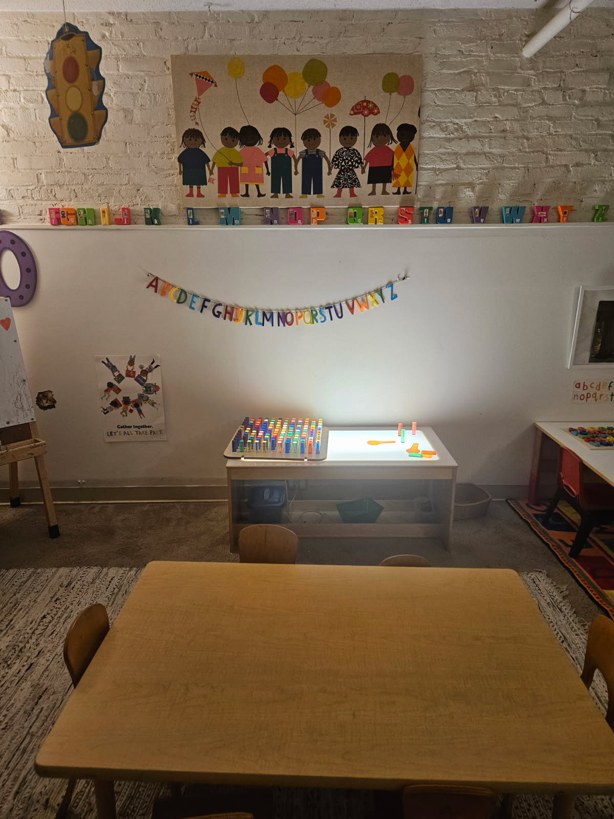 An illuminated light table with translucent pegs, set against a wall of alphabet letters and a hanging alphabet banner.